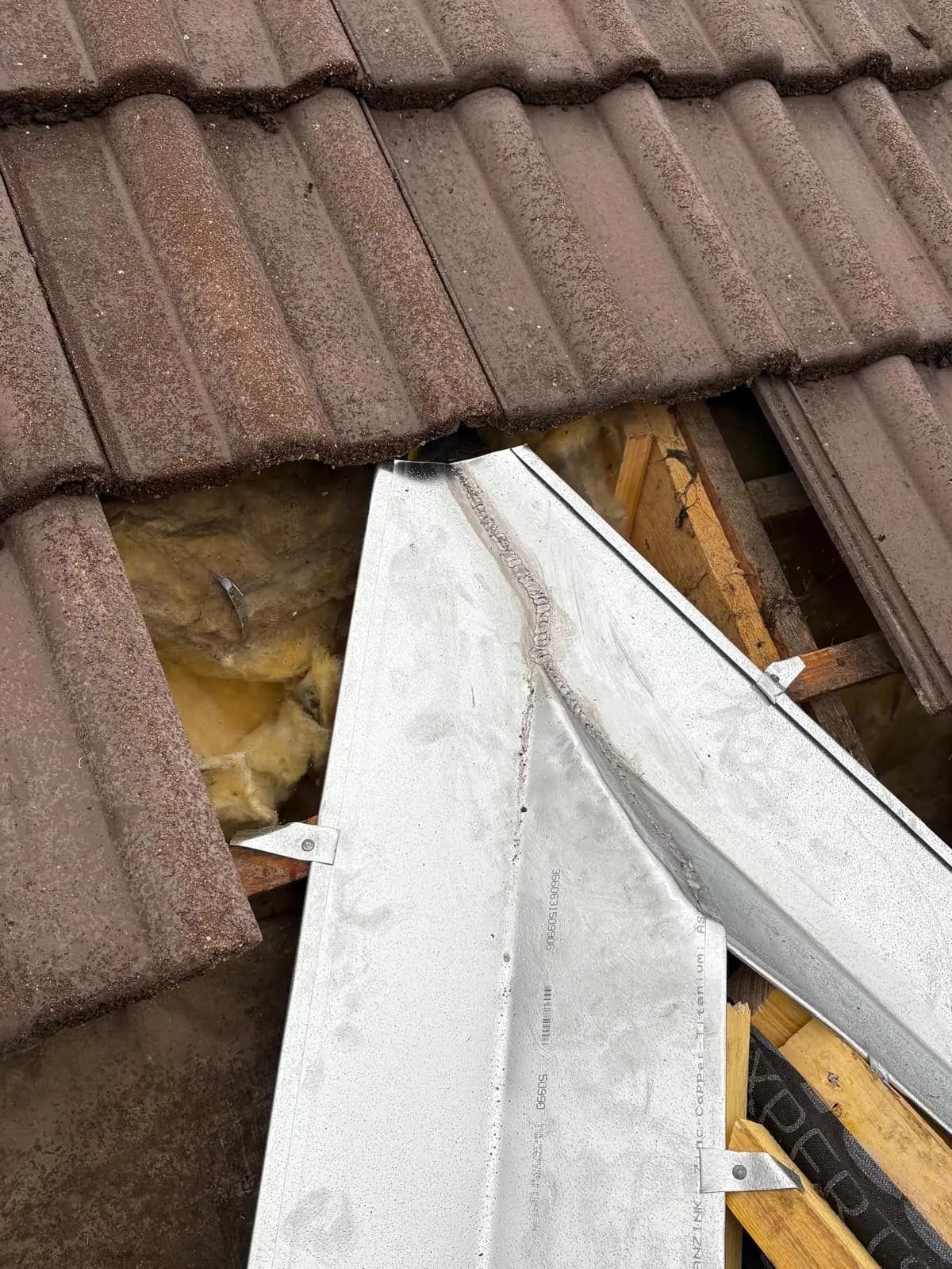Noue en zinc neuve glissée sous les tuiles béton, chevrons et isolant visibles dans l'ouverture — RW Couvreur